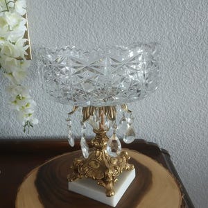 May include: A vintage crystal bowl with a decorative gold-colored metal base and hanging crystal accents. The bowl sits atop a white square base and a round wooden surface. The bowl has a cut glass design.