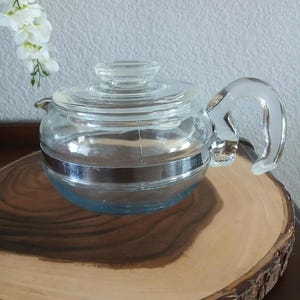 May include: A clear glass teapot with a matching lid and a silver band around the middle. The teapot has a curved handle and sits on a wooden slice. The teapot is designed for brewing tea.