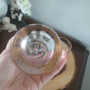 May include: A clear glass teacup with a gold rim and handle. The teacup is held in a hand, with the bottom of the cup facing the viewer. The cup is sitting on a wooden coaster.