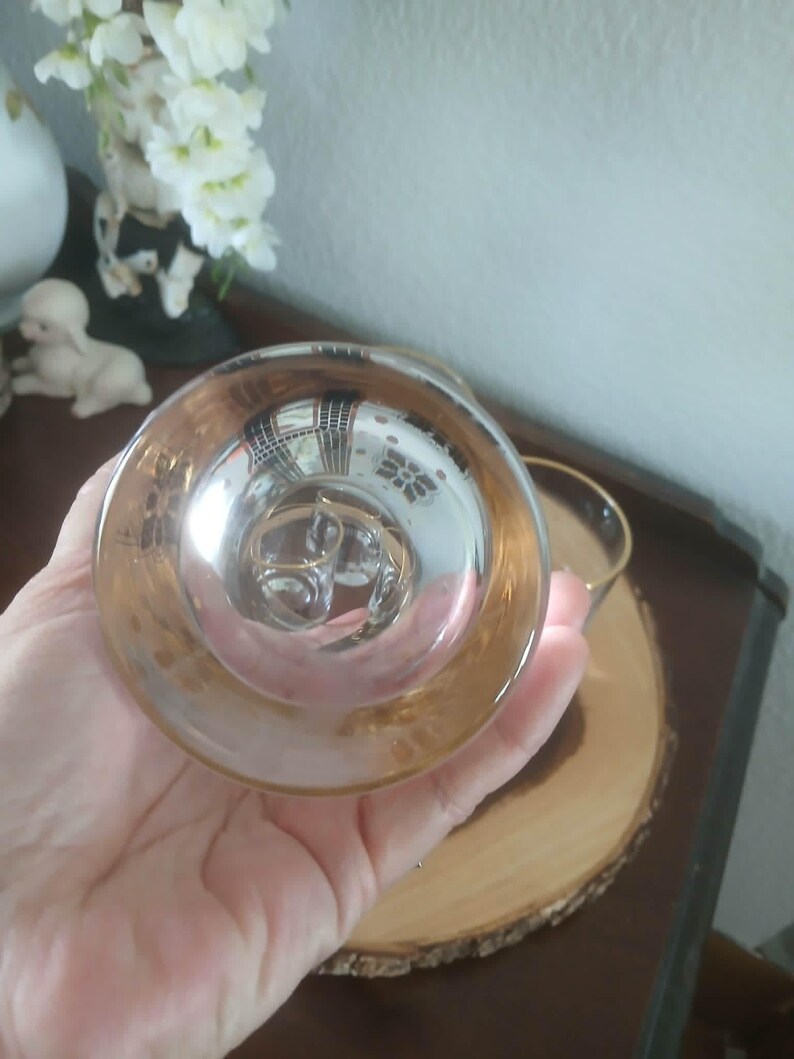 May include: A clear glass teacup with a gold rim and handle. The teacup is held in a hand, with the bottom of the cup facing the viewer. The cup is sitting on a wooden coaster.