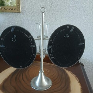May include: A silver-toned metal photo frame with two oval-shaped openings. The frame has a central stem with a small vase and a circular handle at the top. The frame is on a wooden surface.