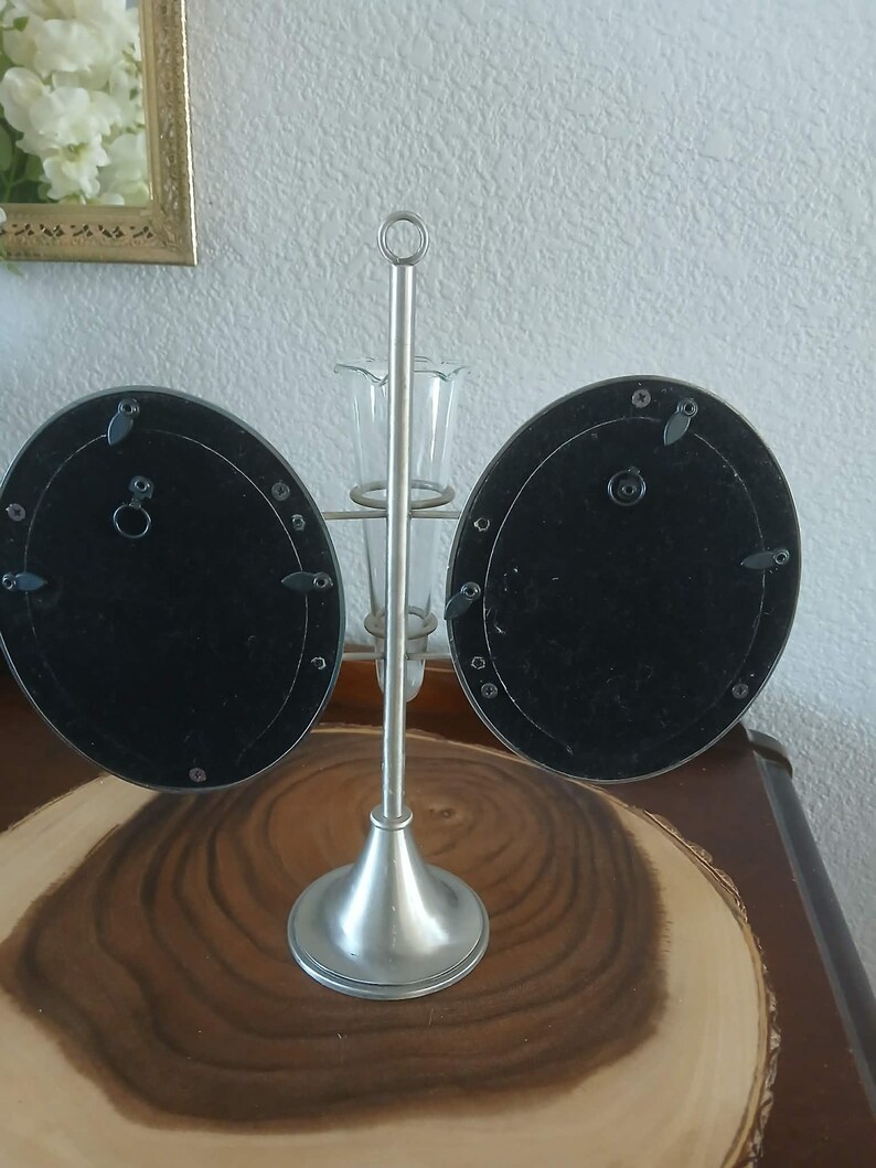 May include: A silver-toned metal photo frame with two oval-shaped openings. The frame has a central stem with a small vase and a circular handle at the top. The frame is on a wooden surface.