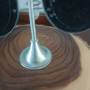 May include: A silver-colored metal display stand with a flared base and a tall, thin stem. The stand is designed to hold a pair of black sunglasses. The stand is set on a wooden surface with a natural grain pattern.