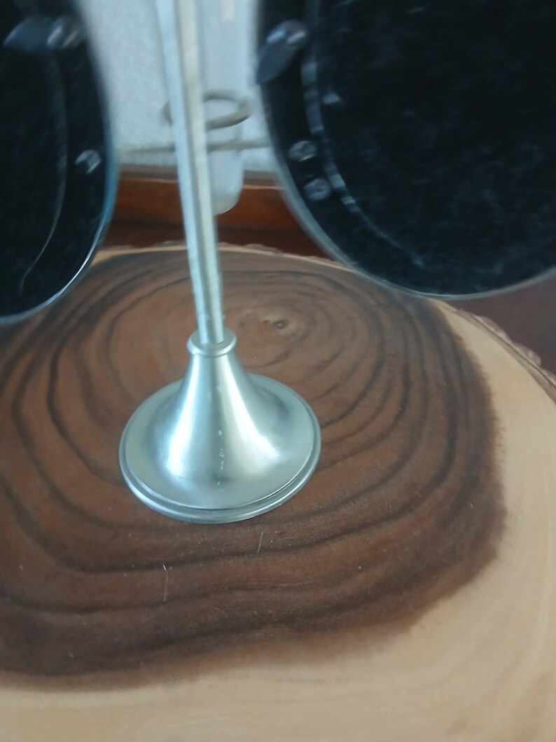May include: A silver-colored metal display stand with a flared base and a tall, thin stem. The stand is designed to hold a pair of black sunglasses. The stand is set on a wooden surface with a natural grain pattern.