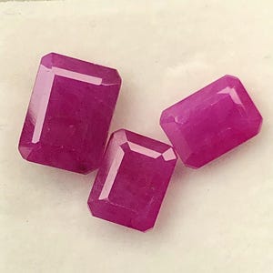 May include: Three faceted, rectangular-cut, deep pink gemstones are displayed in a white plastic container with a cream-colored interior. The gems have a polished, reflective surface, and are arranged in a visually appealing manner.