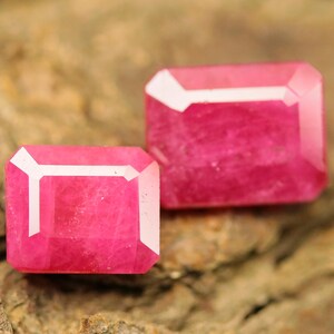 May include: Two rectangular, faceted, pink gemstones with white accents. The gems are displayed on a textured, brown surface, showcasing their vibrant colour and cut. These gems are likely for jewellery making.