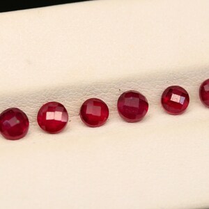 May include: A row of faceted, round, deep red gemstones. The gems are arranged in a line on a white surface, each displaying a brilliant, reflective quality. The gems are likely rubies, known for their rich color and luster.