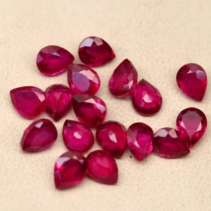 May include: A collection of faceted, teardrop-shaped rubies in varying sizes. The gemstones are a deep, rich red color and are scattered on a light beige surface. The facets catch the light, creating a sparkling effect. These rubies would be suitable for jewelry making.