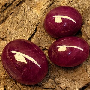 May include: Three polished, oval-shaped, deep red gemstones with a white reflective streak across each. The gems are displayed on a textured, brown, bark-like surface, creating a natural contrast.
