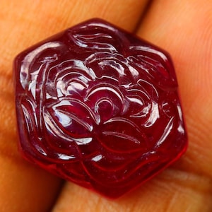 May include: A faceted, hexagonal ruby gemstone with a carved floral design. The deep red stone is held in a hand, showcasing the intricate details of the carved petals and leaves. The gemstone's polished surface reflects light.
