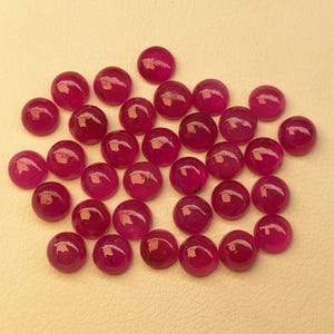 May include: A collection of round, polished ruby gemstones. The gems are a deep, rich red color and have a smooth, reflective surface. The gemstones are scattered across a light beige surface.