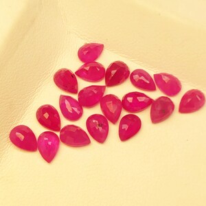 May include: A selection of faceted, teardrop-shaped gemstones in a vivid magenta colour. The gems are displayed on a pale yellow surface, highlighting their polished surfaces and intricate cuts. These gemstones are suitable for jewellery making.