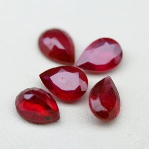 May include: Five loose, pear-shaped rubies with a deep red color and faceted surfaces. Each gemstone displays a polished finish with multiple triangular facets that reflect light, arranged in a cluster on a plain, light-colored surface.