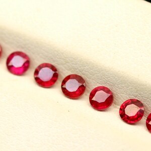 May include: A row of round, faceted red gemstones, possibly rubies, are arranged on a light-colored surface. The gems have a vibrant, deep red color with internal reflections, suggesting a high degree of clarity and polish. The gemstones are evenly spaced.