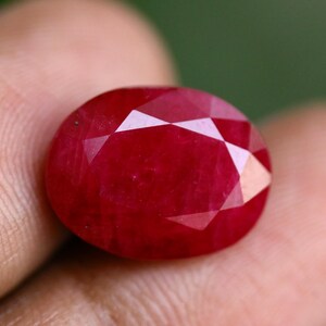 May include: A deep red, oval-shaped gemstone with a faceted surface. The stone displays a rich, saturated crimson hue and a polished finish, featuring multiple triangular and diamond-shaped facets that reflect light across its top surface.