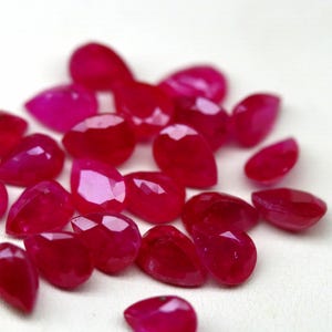 Peut inclure: Un ensemble de pierres précieuses facettées en vrac, d'une teinte rose-rouge profonde et vibrante. Chaque pierre est taillée en forme de goutte ou de poire, avec de multiples facettes qui reflètent la lumière sur leurs surfaces polies.
