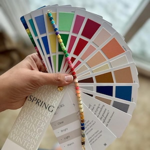 May include: A colour analysis fan deck with a beaded handle, displaying a range of colours. The fan includes cards labelled "SPRING", "SUMMER", "AUTUMN", and "WINTER". The deck also includes text that says "HOUSE OF COLOUR".