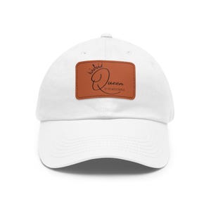 Peut inclure: Casquette de baseball blanche avec un patch en cuir marron. Le patch présente un motif de couronne au-dessus du mot "Queen" en écriture cursive, avec "of the wild things" en dessous. Un accessoire décontracté.