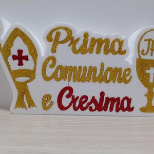 Può includere: Cartello in polistirolo bianco con scritta in glitter dorato "Prima Comunione e Cresima". Il cartello presenta anche una mitra dorata glitterata e un calice dorato glitterato con le lettere "JHS" sopra.