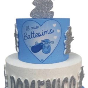 Può includere: Una torta a due piani decorata con una statuina di angelo in glitter argentato, un cuore blu con il testo "Il mio Battesimo" e un paio di scarpine da bebè. Il piano inferiore è bianco con il nome "DOMENICO" in lettere di glitter argentato. La torta è decorata con dettagli in glitter blu e argento.