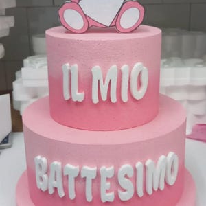 Può includere: Una torta rosa a due piani decorata con glassa bianca. Il piano superiore ha un orsacchiotto rosa che tiene un cuore bianco. Il piano inferiore ha le parole "IL MIO BATTESIMO" in glassa bianca.