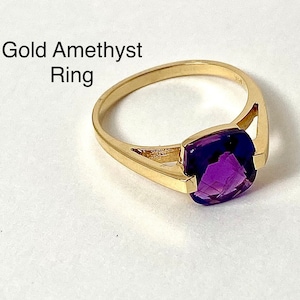 May include: A gold ring with a square-cut, deep purple amethyst gemstone. The ring's band is a simple, polished gold. The text "Gold Amethyst Ring" is displayed in black letters.