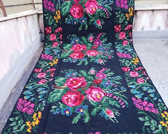 May include: A large, rectangular black rug with a floral design. The rug features a repeating pattern of pink roses, purple and blue flowers, and green leaves. Yellow accents are scattered throughout the design. The rug is displayed outdoors.