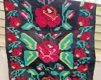 May include: A black rug with a floral design featuring large red roses, green leaves, and accents of pink, blue, and yellow. The rug has a rectangular shape and a woven texture, suitable for home decor.