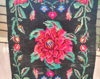 May include: A black wool rug with a floral pattern. The rug features large pink and red flowers with green leaves and stems. The background is black with a subtle texture.