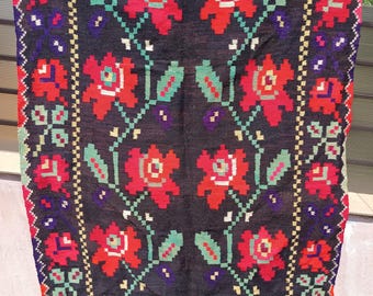 May include: A dark rug with a floral pattern. The rug features large red flowers with green leaves and stems, set against a black background. The border has a repeating geometric design with red, green, and yellow accents. The rug is rectangular.
