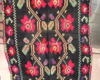 May include: A black woven rug with a floral pattern. The rug features large red flowers with green and yellow accents, arranged symmetrically. Green and purple accents are also present, creating a vibrant design. The rug is hanging outdoors.