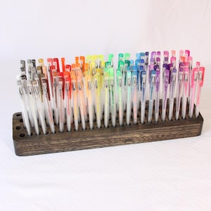 May include: A wooden pen holder with a dark brown stain, holding a rainbow assortment of gel pens. The pens are arranged in rows, with the colors transitioning from brown to purple.