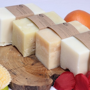 May include: An assortment of handmade soaps in varying shades of cream and beige, wrapped in brown paper. An orange and red rose petals are also visible. A wooden soap dish and a green and yellow brush are in the foreground.