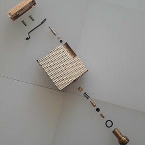 Op de afbeelding: Een gedemonteerde metalen aansteker met een vierkante behuizing en een getextureerd oppervlak met piramidepatroon. De onderdelen omvatten een messingkleurig bovenmechanisme, twee kleine schroeven, een zwarte draadveer, een centrale behuizing.