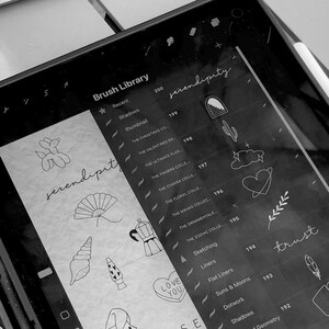 May include: A black and white digital drawing tablet screen displaying a list of brush libraries and icons. The brush libraries include "Shadows", "Thumbnail", "The Christmas Co", "The Valentines Da", "The Ultimate Flash", "The Fingers Colle", "The Chakra Colle", "The Floral Colle", "The Mahas Collec", "The Ornamentals", "The Zodiac Colle", "Sketching", "Liners", "Flat Liners", "Suns & Moons", "Dotwork", "Shadows", and "Geometry". The icons include a balloon animal, a fan, a seashell, a lava lamp, a coffee pot, a heart with the words "Love You", a cactus, a cloud with a heart, a branch with leaves, and a feather.