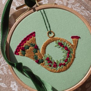 May include: Embroidered Christmas horn ornament in a wooden hoop. The horn is gold with red and green accents, and a green velvet ribbon is attached for hanging. The background is a light green fabric.