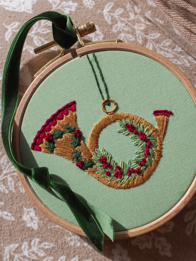 May include: Embroidered Christmas horn ornament in a wooden hoop. The horn is gold with red and green accents, and a green velvet ribbon is attached for hanging. The background is a light green fabric.