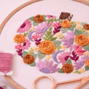 Op de afbeelding: Geborduurd pompoenontwerp in een houten hoepel. De pompoen is gevuld met oranje, roze en paarse bloemen, groene bladeren en een bruine stengel. Roze en groen borduurgaren en ros&eacute;gouden schaar zijn ook in de afbeelding te zien.