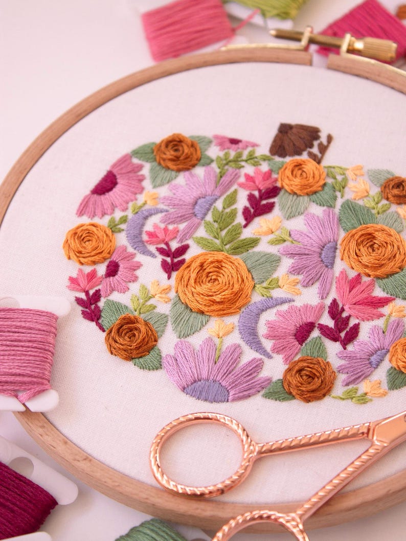 Op de afbeelding: Geborduurd pompoenontwerp in een houten hoepel. De pompoen is gevuld met oranje, roze en paarse bloemen, groene bladeren en een bruine stengel. Roze en groen borduurgaren en ros&eacute;gouden schaar zijn ook in de afbeelding te zien.