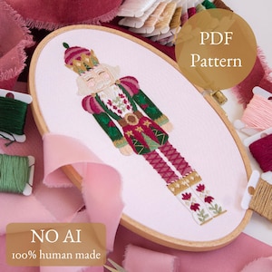 May include: An embroidered design of a Nutcracker soldier in a wooden embroidery hoop. The Nutcracker is dressed in a red, green, and gold uniform with a crown. The text "PDF Pattern" and "NO AI 100% human made" are also visible.