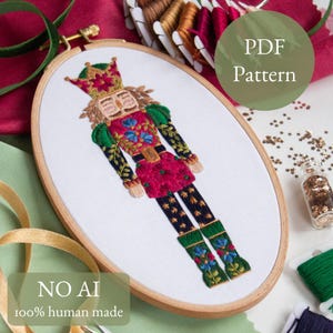 May include: An embroidered nutcracker design in a wooden hoop. The artwork features a red and green uniform with floral accents, a gold crown, and black trousers with star details. Text reads "PDF Pattern" and "NO AI 100% human made".