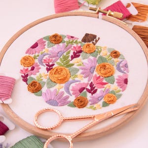 Op de afbeelding: Een borduurring met een bloemenpompoenontwerp. De pompoen is gevuld met oranje, roze en paarse bloemen en groene bladeren. Koperkleurige scharen en spoelen garen in verschillende kleuren omringen de ring, wat een ambachtelijk thema cre&euml;ert.