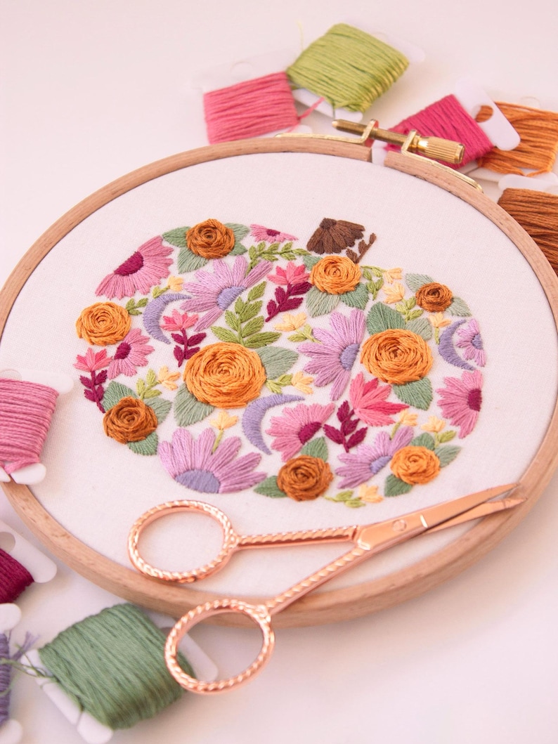 Op de afbeelding: Een borduurring met een bloemenpompoenontwerp. De pompoen is gevuld met oranje, roze en paarse bloemen en groene bladeren. Koperkleurige scharen en spoelen garen in verschillende kleuren omringen de ring, wat een ambachtelijk thema cre&euml;ert.