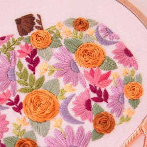 Op de afbeelding: Geborduurd pompoenontwerp in een houten hoepel. De pompoen is gevuld met oranje, roze, paarse en groene bloemborduurwerk. Een gouden schaar ligt in de rechter benedenhoek. De borduursels zijn op een witte stof.