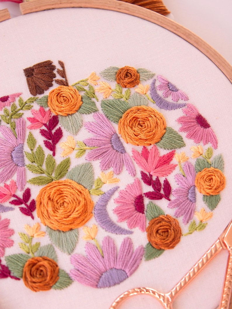 Op de afbeelding: Geborduurd pompoenontwerp in een houten hoepel. De pompoen is gevuld met oranje, roze, paarse en groene bloemborduurwerk. Een gouden schaar ligt in de rechter benedenhoek. De borduursels zijn op een witte stof.