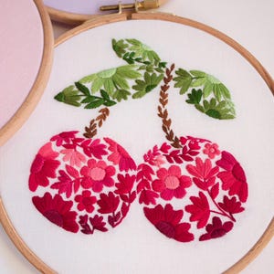 May include: Embroidered cherry design on white fabric stretched within a wooden hoop. The cherries are filled with red and pink floral patterns, with green leaves and brown stems. Other floral embroidery hoops are visible in the background.