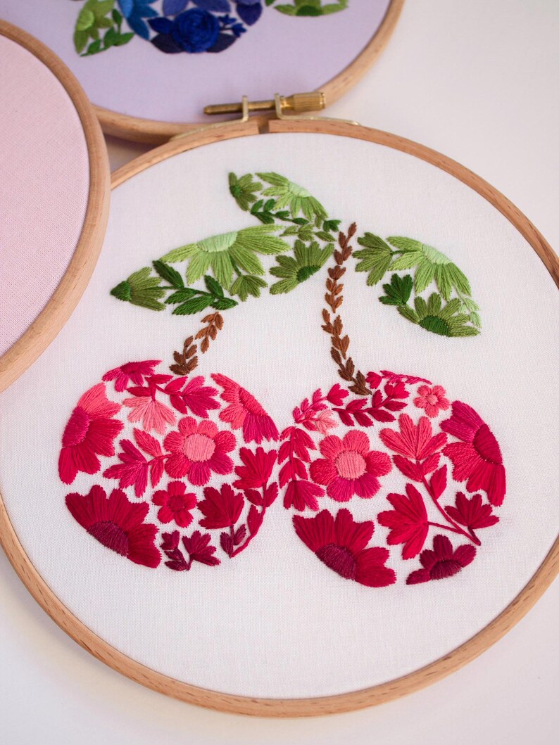 May include: Embroidered cherry design on white fabric stretched within a wooden hoop. The cherries are filled with red and pink floral patterns, with green leaves and brown stems. Other floral embroidery hoops are visible in the background.