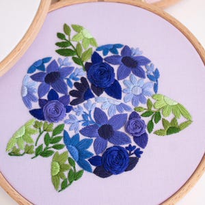May include: Embroidered floral design in shades of blue and green on a lavender fabric, set in a wooden embroidery hoop. The design features various flower types and leaves, creating a detailed and textured artwork.