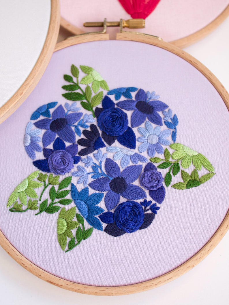 May include: Embroidered floral design in shades of blue and green on a lavender fabric, set in a wooden embroidery hoop. The design features various flower types and leaves, creating a detailed and textured artwork.
