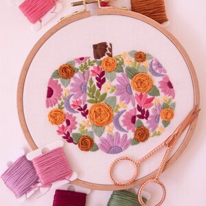 Op de afbeelding: Een borduurring met een bloemenpompoenontwerp in tinten oranje, roze, paars en groen. Spoelen borduurgaren in diverse kleuren en een gouden schaar zijn rond de ring geplaatst.